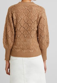 Maglione lavorato a maglia in marrone chiaro con un motivo a diamante e finitura testurizzata; presenta polsini e orlo a costine, indossato sopra una gonna bianca.