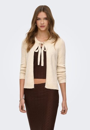 Mlada ženska z dolgimi lasmi, oblečena v kremasto kratko jopico, ki je zavezana pri vratu, čez teksturiran rjav top in ujemajočo se krilo.