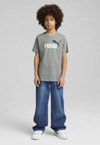 Šedé tričko s bílým logem "PUMA", doplněné volnými modrými džínami. Modelka nosí bílé tenisky a stojí s rukama podél těla.