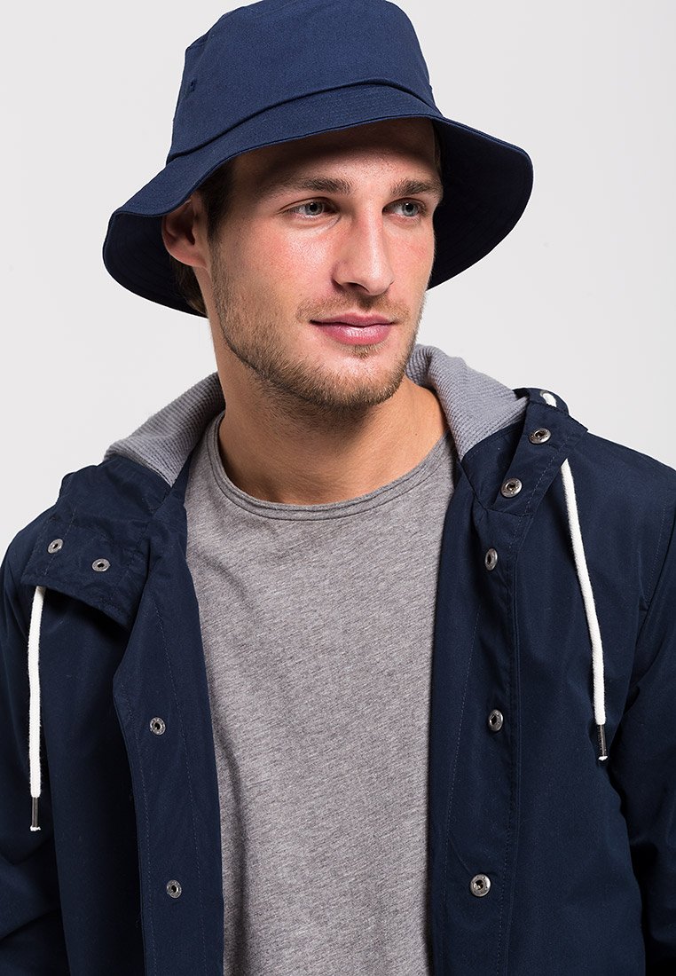 Navyblauer Buckethat mit breiter Krempe, aus strapazierfähigem Stoff gefertigt. Getragen mit einem grauen T-Shirt und einer geöffneten navyblauen Jacke mit silbernen Details.