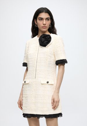 Hosszú, sötét hajú nő texturált, krémszínű, rövid ujjú ruhát visel fekete fodros szegéllyel és nagy fekete virágdísszel a gallér közelében.