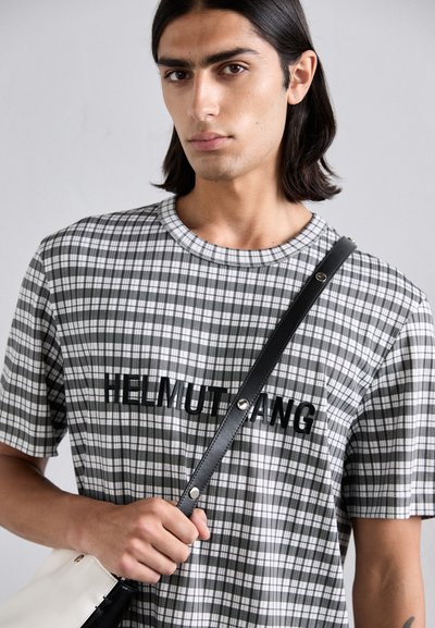 T-shirt à carreaux gris et blanc avec un logo noir, col rond, manches courtes ; modèle tenant un sac à bandoulière noir avec un corps blanc contrastant.