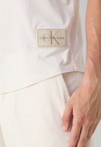 Vit bomullst-shirt med en rektangulär beige etikett med präglad "Calvin Klein Jeans"-logotyp och "CK"-initialer, kombinerad med ljusa byxor.