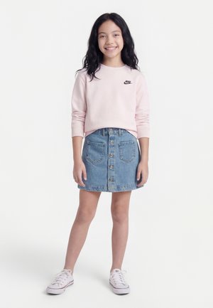 Lachend meisje met een lichtroze Nike-sweatshirt, een knoopjes spijkerrok en witte sneakers, staand tegen een effen witte achtergrond.