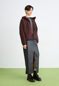 Chaqueta de forro polar marrón con capucha, camiseta rayada, falda de cuero negra con abertura frontal y botas negras con cordones y suelas gruesas. Suelo verde.