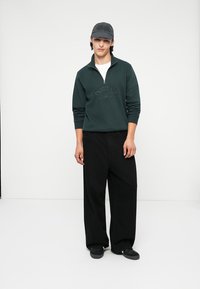 Dark green zip-up sweatshirt with embroidered "ORIGINALS" text, paired with loose black trousers and a dark gray cap.