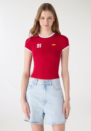Giovane donna che indossa una maglietta aderente rossa a maniche corte con rifiniture bianche, numero 91 e piccolo dettaglio con bandiera, abbinata a pantaloncini di jeans azzurri.