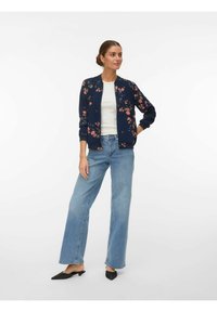Femme debout portant une veste bomber bleue à fleurs, un haut blanc, un jean bleu clair à jambes larges et des escarpins noirs à talons bas et bouts pointus sur fond blanc.