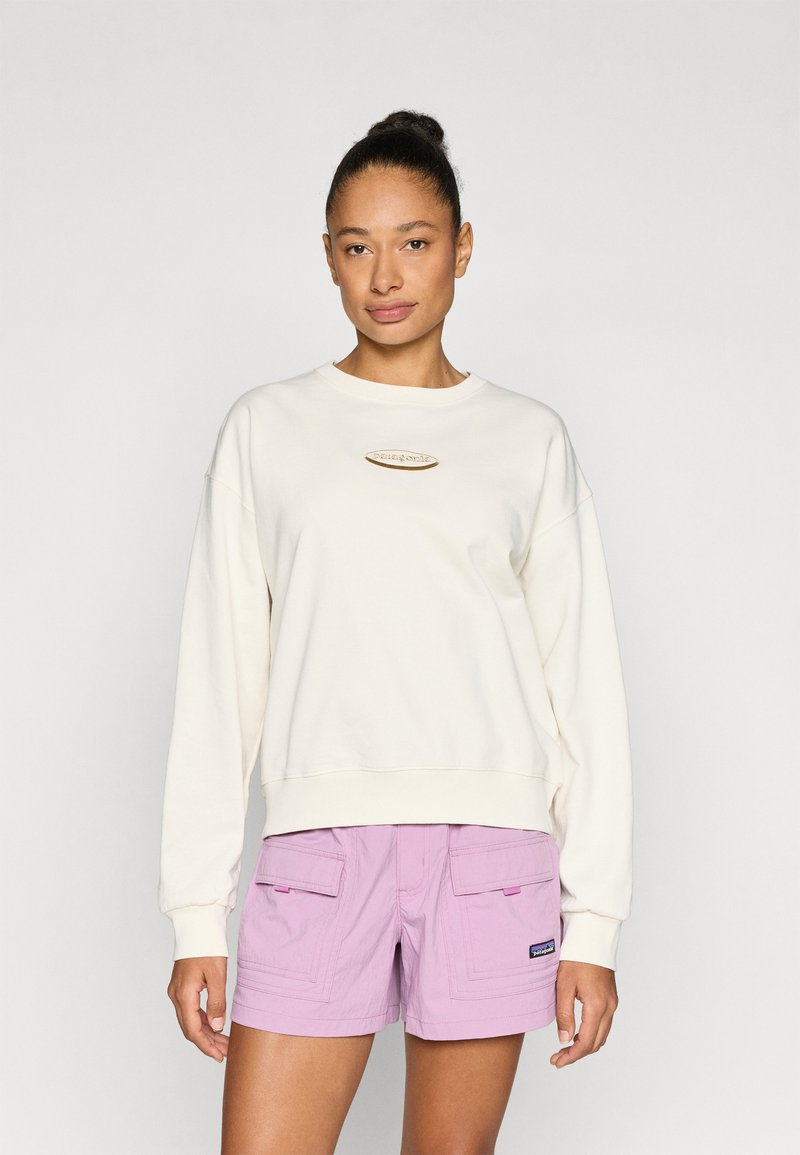 Εκρού φούτερ με στρογγυλή λαιμόκοψη και λογότυπο σε χρυσό τόνο, συνδυασμένο με ανοιχτό μωβ cargo σορτς που διαθέτει εμπρόσθιες τσέπες και ραφές.