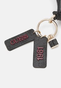 Porte-clés noir texturé avec anneau doré, deux étiquettes portant les inscriptions "GUESS" et "1981" en rouge foncé, et un petit pendentif cadenas noir et doré.