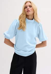 Felpa azzurro chiaro con colletto alto, maniche a sbuffo e vestibilità rilassata. Materiale morbido e dettaglio a coste sull'orlo. Abbinata a pantaloni neri.