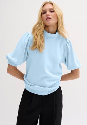 Lichtblauwe sweatshirt met een hoge kraag, opbollende mouwen en een relaxed fit. Zacht materiaal en geribbelde zoomdetails. Gecombineerd met zwarte broek.