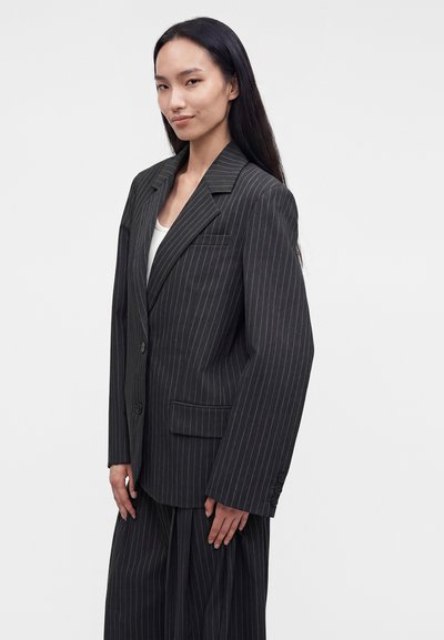 Femme aux longs cheveux foncés porte un costume noir à rayures et un haut blanc, debout devant un fond uni clair, face à l'avant.