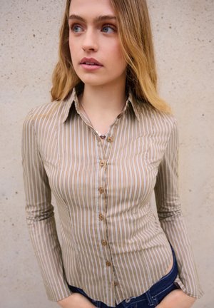 Tânără cu părul lung și blond purtând o cămașă cu dungi bej și albe și blugi albaștri, stând în picioare în fața unui perete bej.