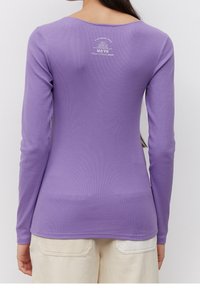 Lila långärmad tröja i ribbat tyg, med en rundad rygg och en liten vit logotypdetalj på övre ryggen. Avskuren fåll med åtsittande passform.