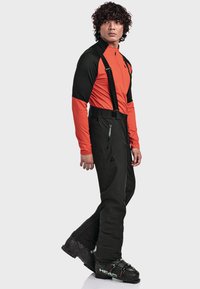 Mann in schwarzen Skihosen mit Trägern, einer orange-schwarzen Jacke und schwarzen Skischuhen, der vor einem weißen Hintergrund steht.