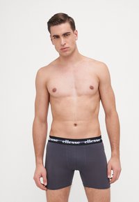 Graue Boxer Shorts mit einem schwarzen elastischen Bund, der mit weißem "ellesse"-Branding versehen ist. Glatte Textur, figurbetonter Schnitt und Oberschenkellänge.