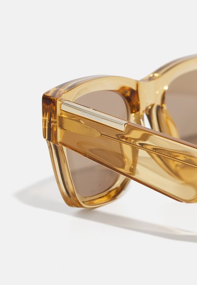 Braungetönte Sonnenbrille mit transparenten, bernsteinfarbenen Fassungen. Verfügt über ein klobiges Design, goldfarbene Metallakzente und ein auffälliges Logo am Bügel.