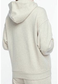 Osoba v volné, světle béžové mikině s kapucí, zobrazená zespodu, v kombinaci s odpovídajícími světle béžovými kalhotami.