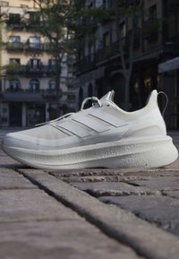 Valkoinen Adidas-juoksukenkä, jossa on kolme raitaa, mukulakivikadulla kaupunkimaisemassa, taustalla rakennuksia ja puita.