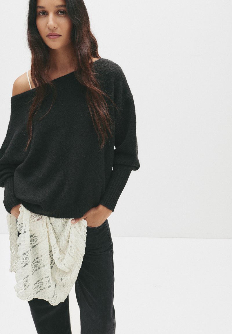 Zwarte, loszittende trui met textuurgebreid uiterlijk en een off-the-shoulder ontwerp. Gecombineerd met zwarte jeans en een witte kanten sjaal die casual is gedrapeerd.