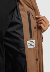 Braune Outdoorjacke mit schwarzem Futter, ausgestattet mit einer Reißverschlusstasche und einem Stoffetikett mit der Aufschrift "Eine warme Umarmung aus Stoff."