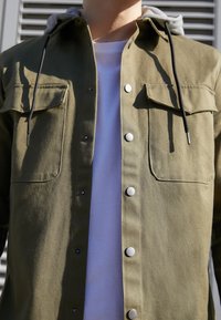 Persona vistiendo una chaqueta de botón color verde oliva con bolsillos en el pecho y cordones negros sobre una camiseta blanca.