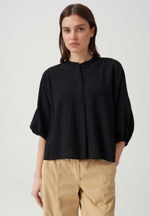 Femme aux cheveux bruns mi-longs porte un chemisier noir ample à manches trois-quarts et un pantalon beige sur un fond uni.
