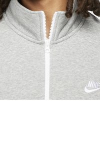 Detailní záběr na šedou mikinu na zip s bílým zipem a bílým logem Nike na hrudi, kterou má na sobě osoba s kudrnatými vlasy.