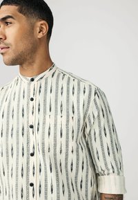 Chemise beige clair avec des motifs géométriques noirs, col montant, sept boutons et une seule poche poitrine. Le tissu semble texturé.
