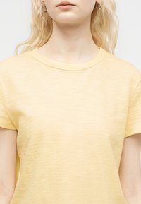 Νεαρή γυναίκα με μακριά ξανθά μαλλιά, που φοράει ανοιχτό κίτρινο t-shirt με κοντά μανίκια και στρογγυλή λαιμόκοψη, καθώς και ασημένια σκουλαρίκια κρίκους, φωτογραφημένη από τη μέση και πάνω.