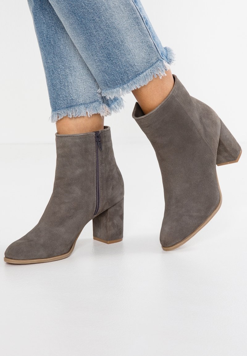 Bottes cheville en daim gris avec bout pointu et talon épais. Détail de fermeture éclair latérale et surface légèrement texturée. Portées avec un jean à ourlet effiloché.