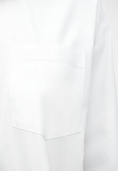 Witte katoenen shirtstof met een close-up van een rechthoekige borstzak die aan de linkerkant is gestikt.