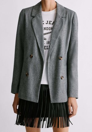 Femme portant un blazer gris croisé sur un t-shirt blanc à motif et une jupe en cuir noire à franges, debout devant un fond uni.