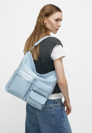 Młoda kobieta z długimi włosami, ubrana w szarą bezrękawnik i dżinsy, niesie dużą jasnoniebieską skórzaną torbę z trzema przednimi kieszeniami.