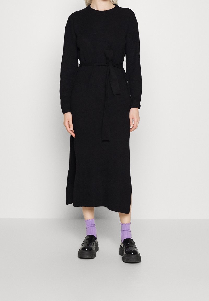 Femme portant une longue robe en tricot noire avec une ceinture à la taille, associée à des chaussettes violettes et des mocassins noirs épais, debout contre un mur uni.