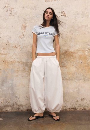 Žena s dlhými tmavými vlasmi, oblečená v svetlomodré tričko s nápisom "Argentina", voľné biele nohavice a čierne žabky, stojaca pri textúrovanej stene.