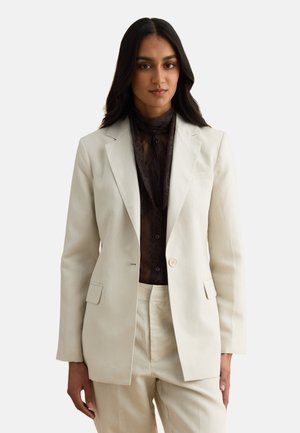 Femme portant un costume sur mesure crème avec un blazer boutonné à un seul bouton et un pantalon crème, associé à un haut en dentelle noire à col montant.