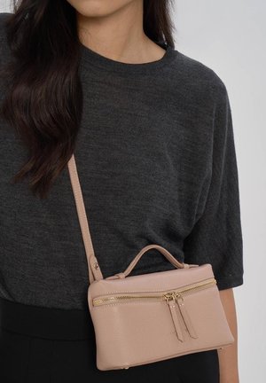 Kobieta ubrana w ciemnoszary top i czarne spodnie, z beżową skórzaną torbą na ramię z złotym zamkiem błyskawicznym i podwójnymi suwakami.