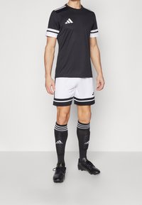 Black short-sleeve soccer jersey with white accents, paired with white shorts featuring black stripes and knee-length black socks. Black cleats.