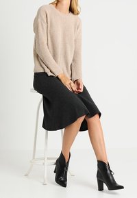 Beige resoitetu neule, jossa roikkuvat olkapäät, yhdistetty tumman polvenpituiseen hameeseen ja mustiin nilkkureihin, joissa on sivuvetoketjut.