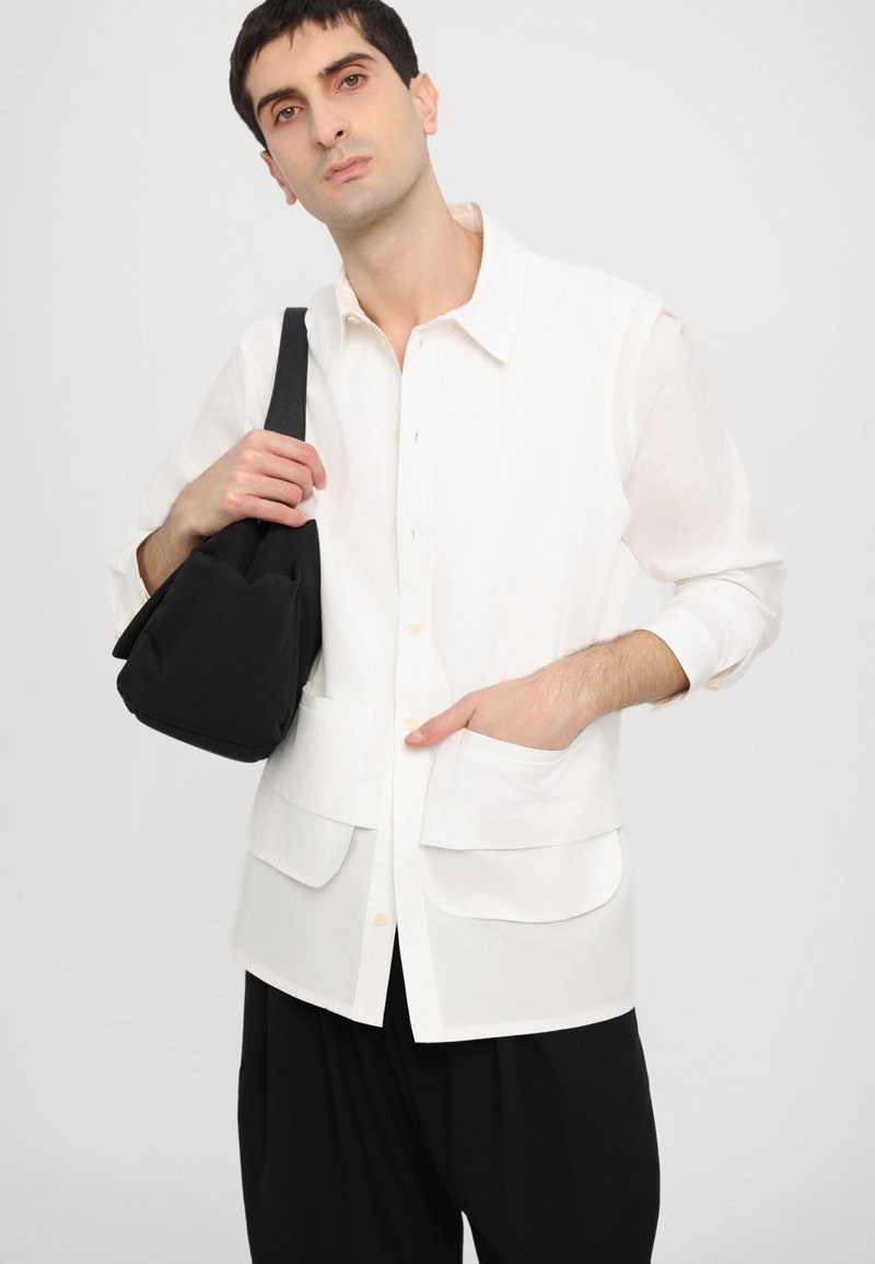 Camicia bianca con bottoni e ampie tasche frontali, abbinata a una borsa a spalla nera in tessuto. Il modello indossa pantaloni neri larghi. Design semplice e pulito.