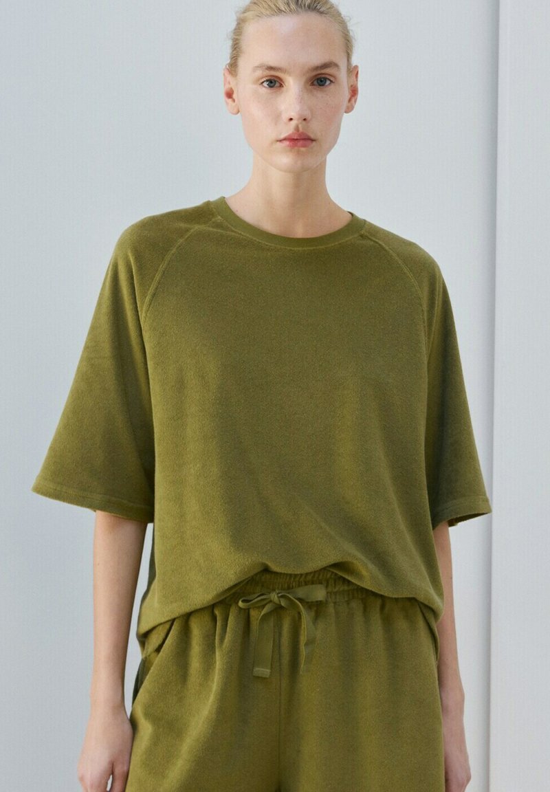Femme à la peau claire et aux cheveux blonds portant un haut ample à manches courtes vert olive et un pantalon assorti à cordon, debout devant un fond clair.