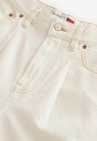Jeans en denim couleur crème avec une coupe droite, dotés de quincaillerie en cuivre, de surpiqûres ton sur ton et de coutures latérales visibles pour la structure.