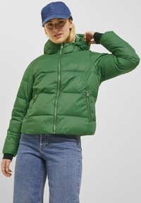 Grön vadderad jacka med en kortad design, som har en hög krage, dragkedja framtill och två dragkedjefickor. Bärs med blå jeans och en denimkeps.