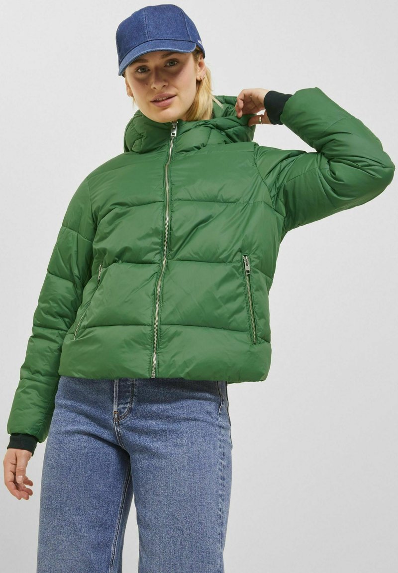 Grön vadderad jacka med en kortad design, som har en hög krage, dragkedja framtill och två dragkedjefickor. Bärs med blå jeans och en denimkeps.