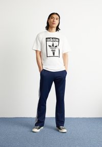 White cotton T-shirt with a black Adidas logo featuring a torch and flames. Paired with navy blue pants and black sneakers with white soles.