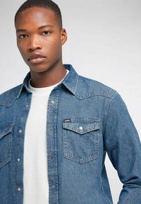 Camicia di jeans di colore azzurro chiaro con due tasche sul petto, bottoni a pressione e un'etichetta sulla tasca sinistra. Indossata sopra una maglietta bianca a girocollo.