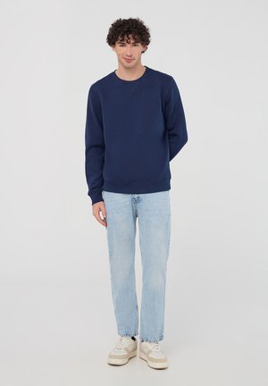 Felpa blu navy con scollatura a girocollo, maniche lunghe e polsini a costine, abbinata a jeans azzurri a gamba dritta e sneakers bianche.