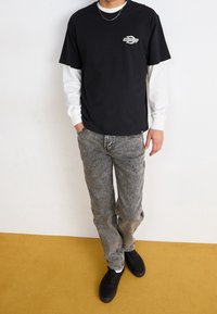 Černé bavlněné tričko přes bílé tričko s dlouhým rukávem, doplněné šedými vzorovanými džínami a černými teniskami. Viditelný náhrdelník jako doplněk.